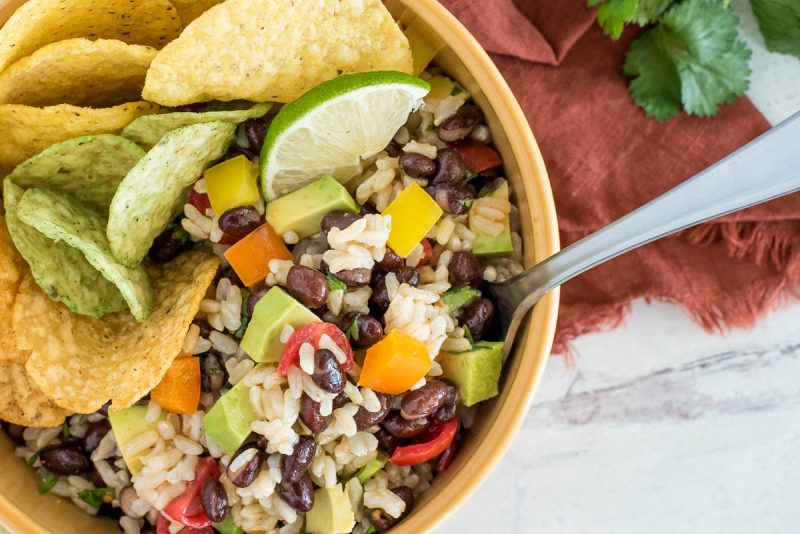 close up on a bowl of brown rice and black bean salad wth tortilla chips and lime